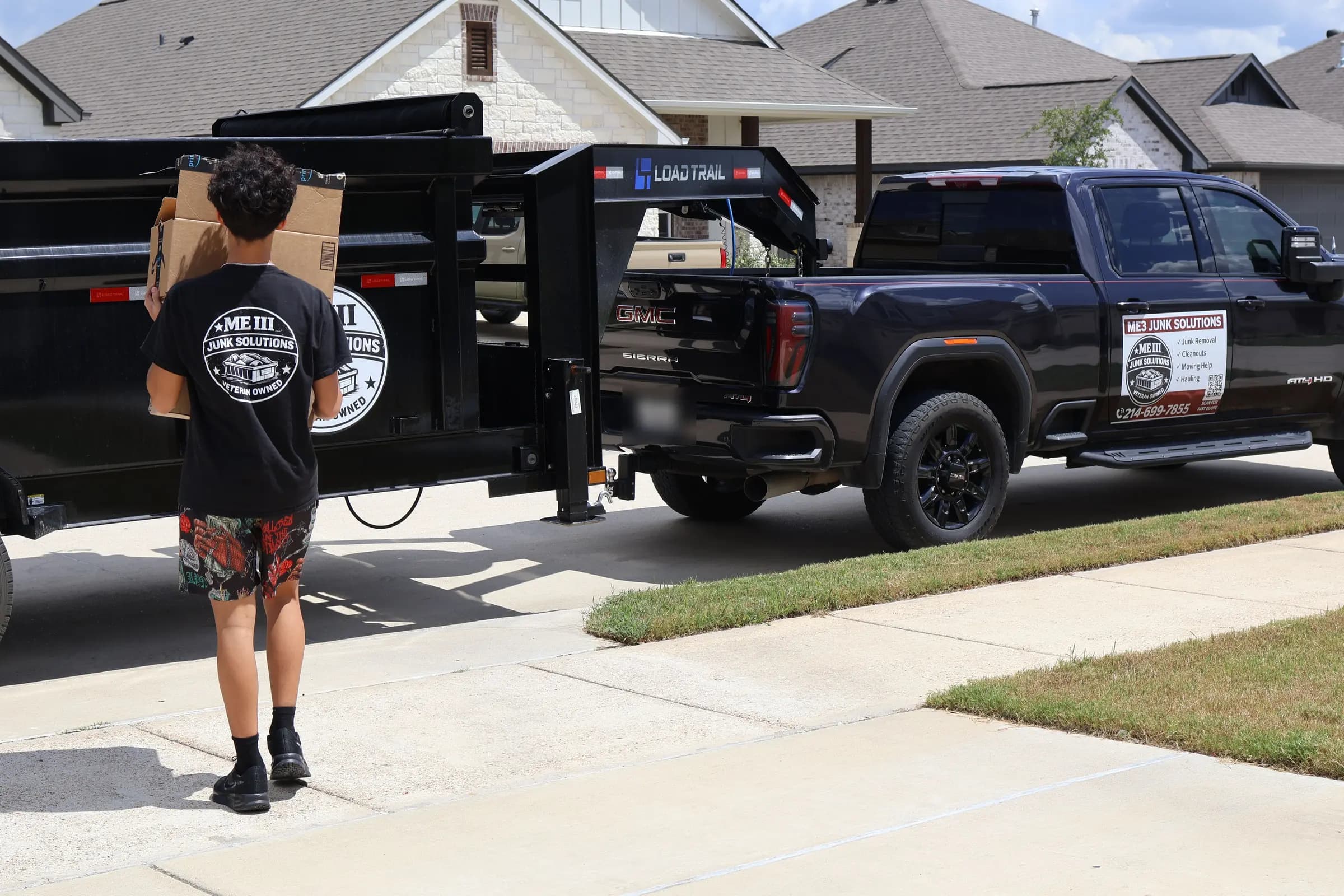 ME3 Junk Solutions pickup and dump trailer parked in a driveway, with a crew member carrying a box to the trailer