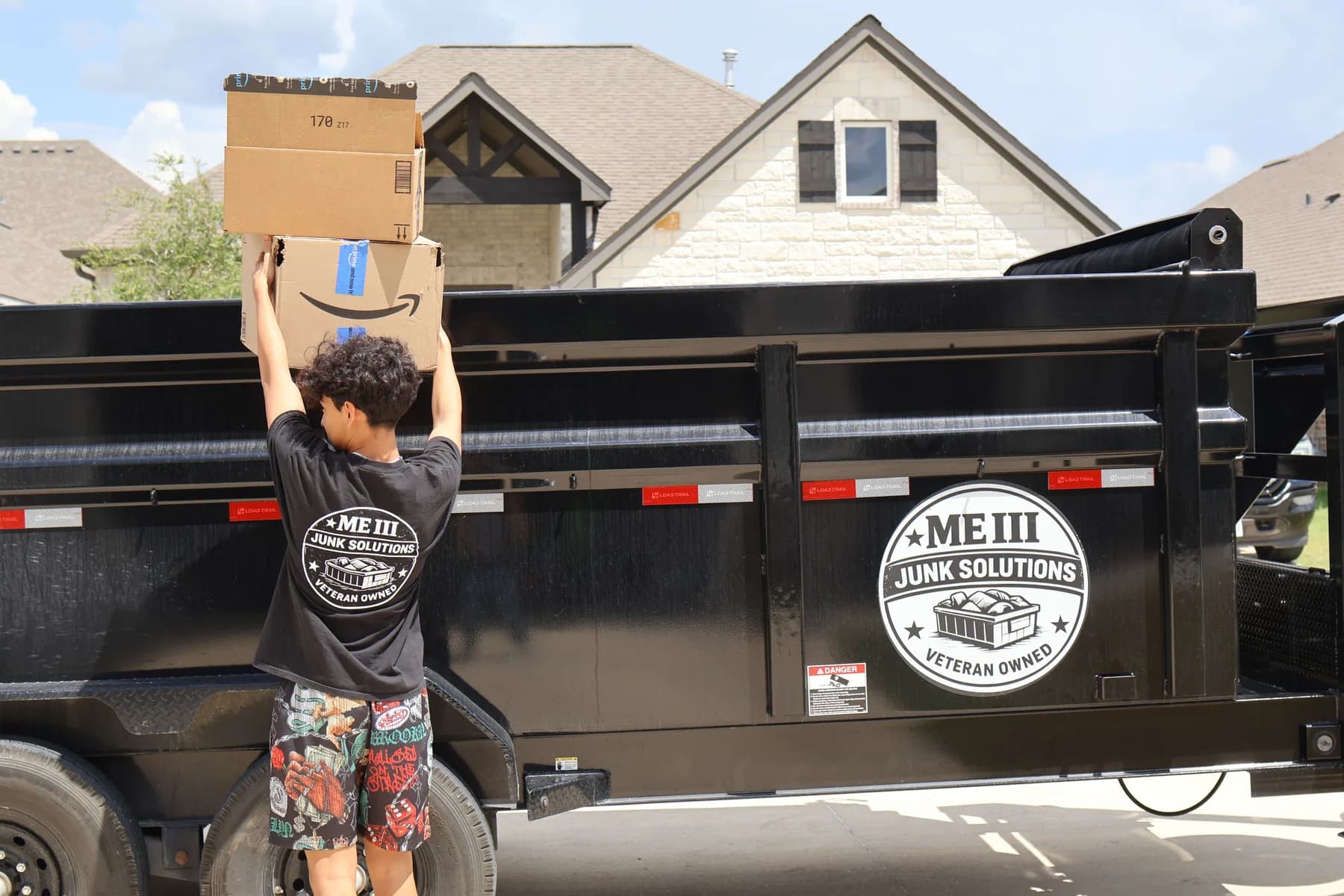An ME3 Junk Solutions crew member lifting boxes into the black dump trailer outside a house